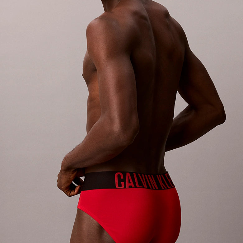 A man shown from the back wearing bright red Calvin Klein briefs with a black waistband and red logo text, against a plain light gray studio background.