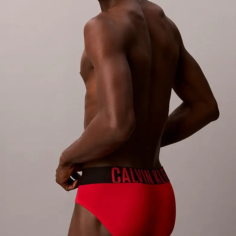 A man shown from the back wearing bright red Calvin Klein briefs with a black waistband and red logo text, against a plain light gray studio background.