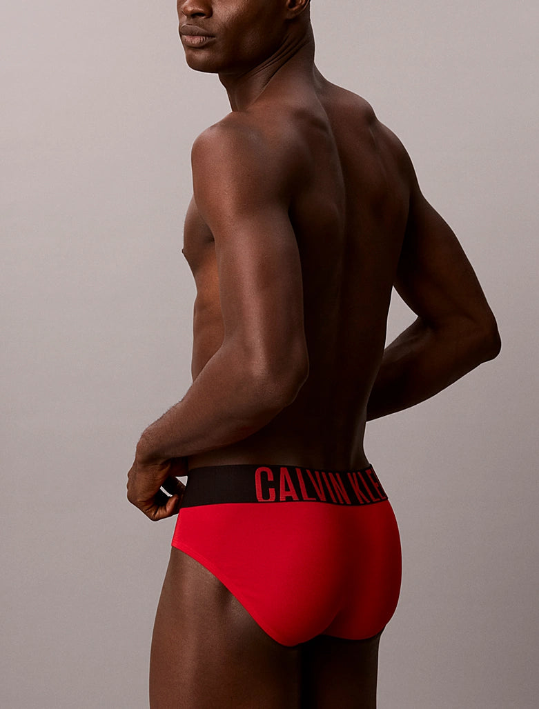 A man shown from the back wearing bright red Calvin Klein briefs with a black waistband and red logo text, against a plain light gray studio background.