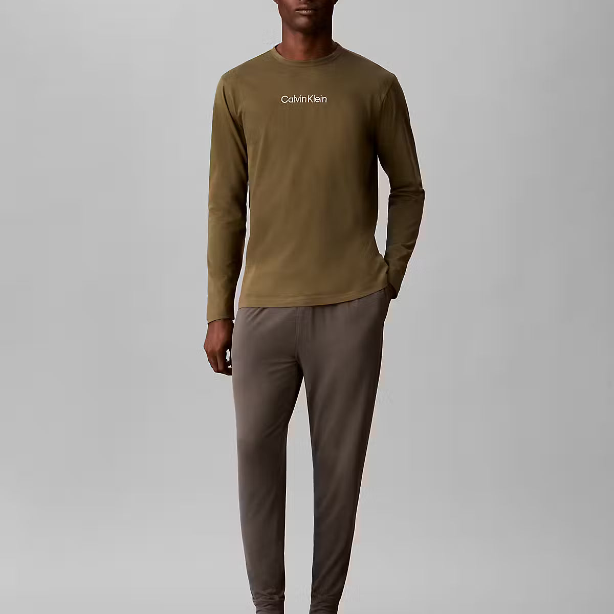 A man modeling a two-piece Calvin Klein loungewear set consisting of an olive green long-sleeve shirt and dark brown pants, standing against a neutral gray studio background.