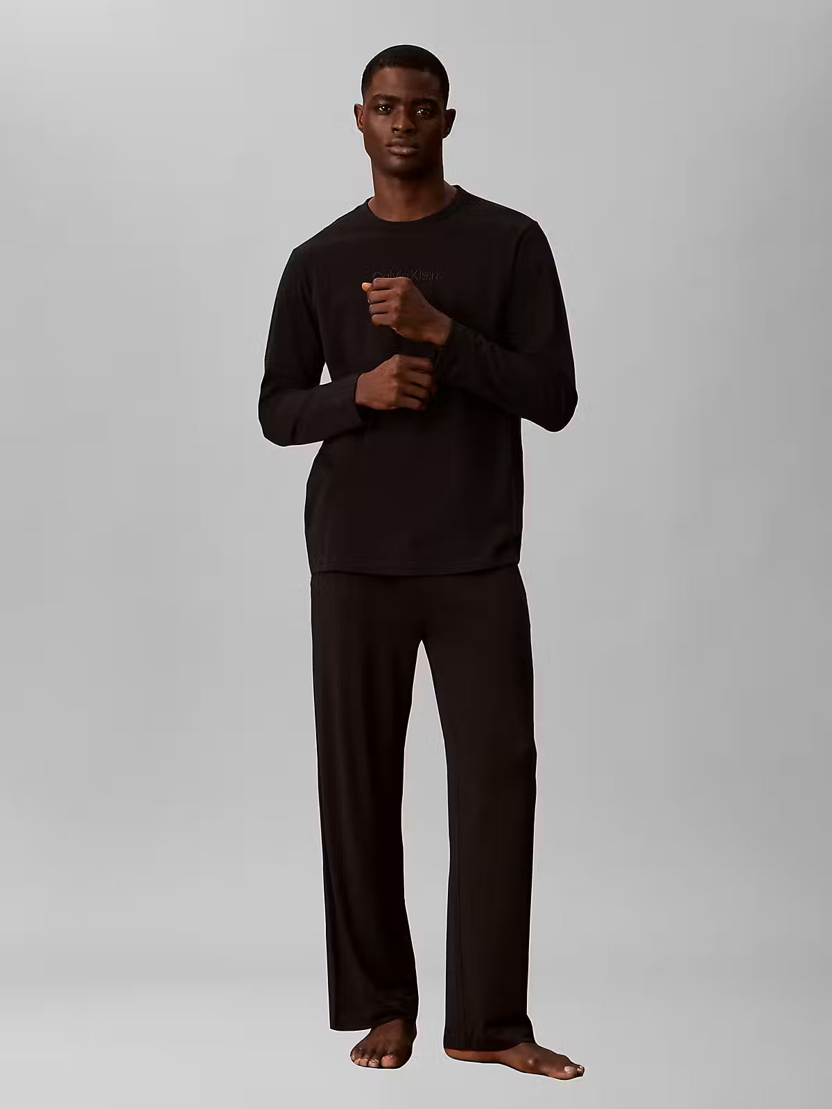 A man modeling a matching black long-sleeve shirt and wide-leg pants lounge set against a solid light gray background.