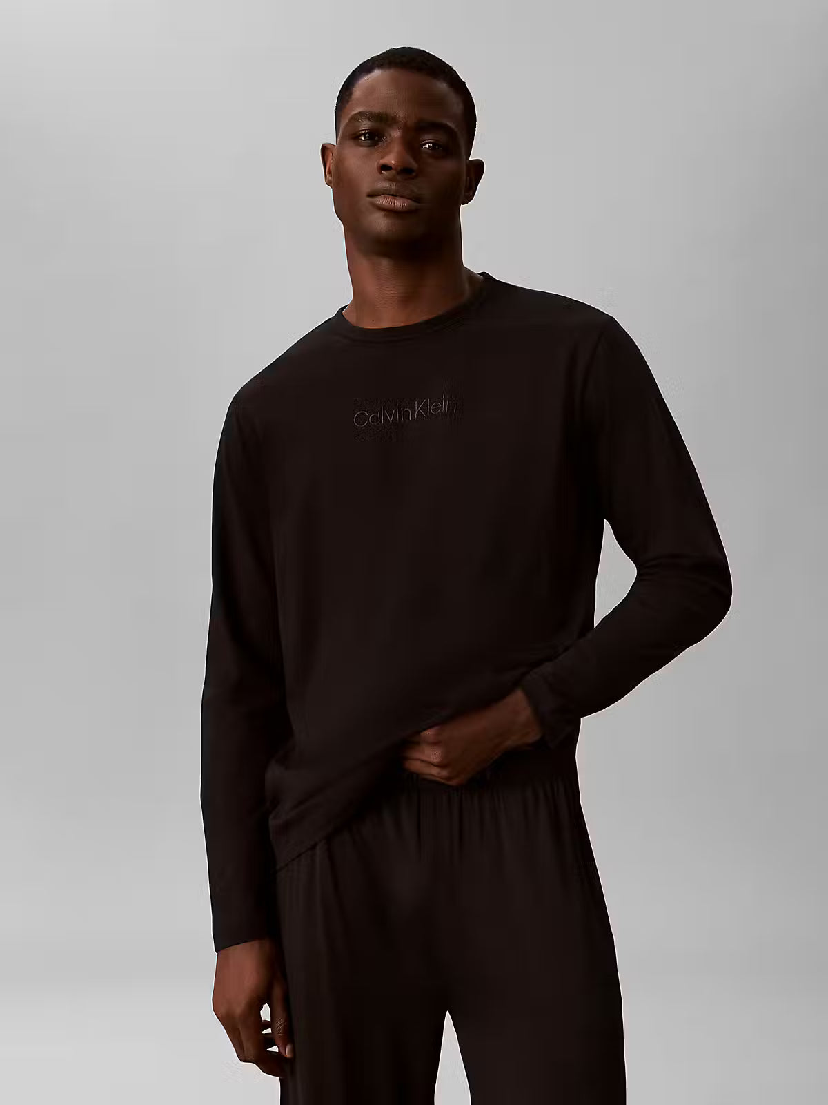 A man modeling a black long-sleeve crewneck sweatshirt with a subtle tonal Calvin Klein logo on the chest, paired with matching black sweatpants, against a neutral gray studio background.