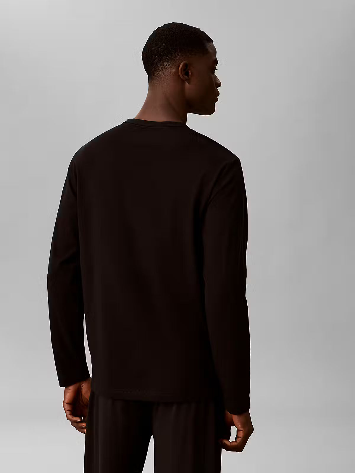 A man shown from the back wearing a dark brown long-sleeve crewneck shirt and matching dark pants against a light gray studio background.
