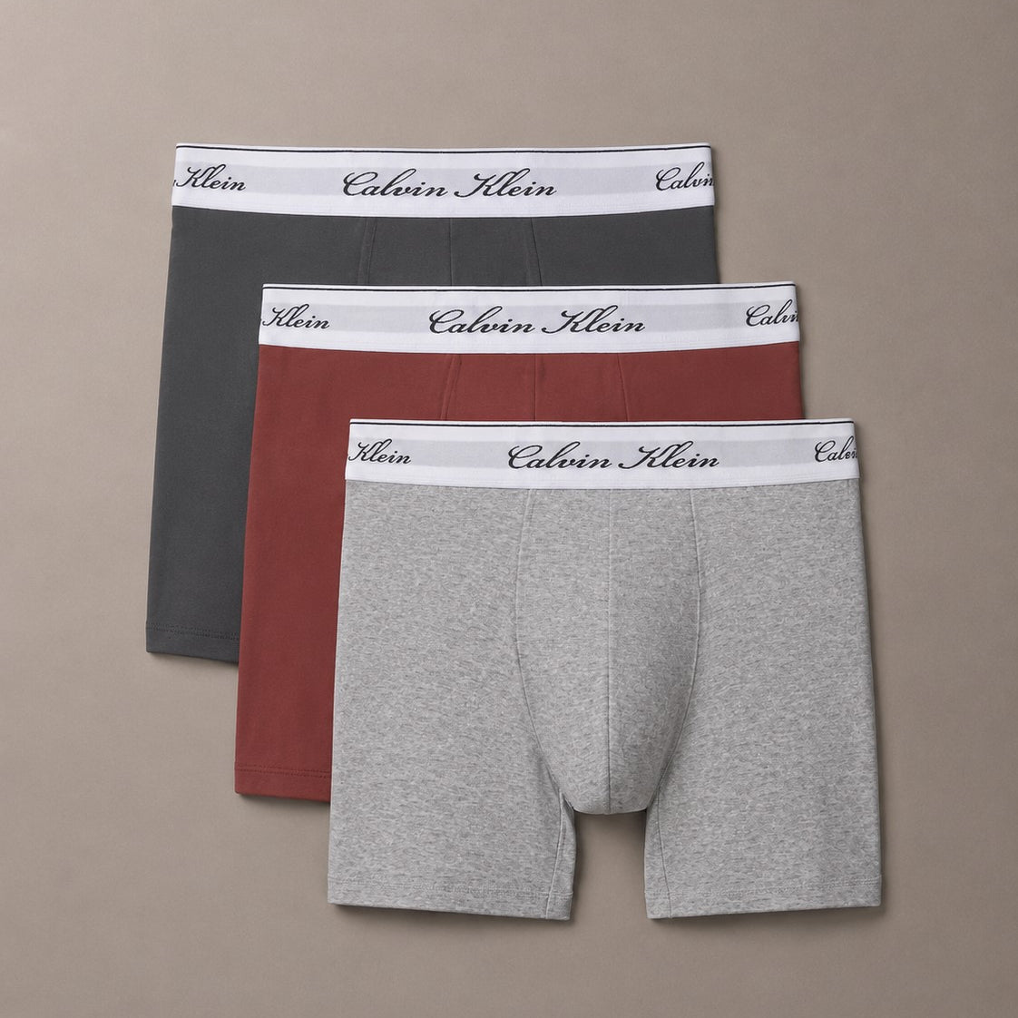Three pairs of Calvin Klein men's boxer briefs in dark gray, rust red, and light heather gray, arranged in a fan shape on a solid taupe background.