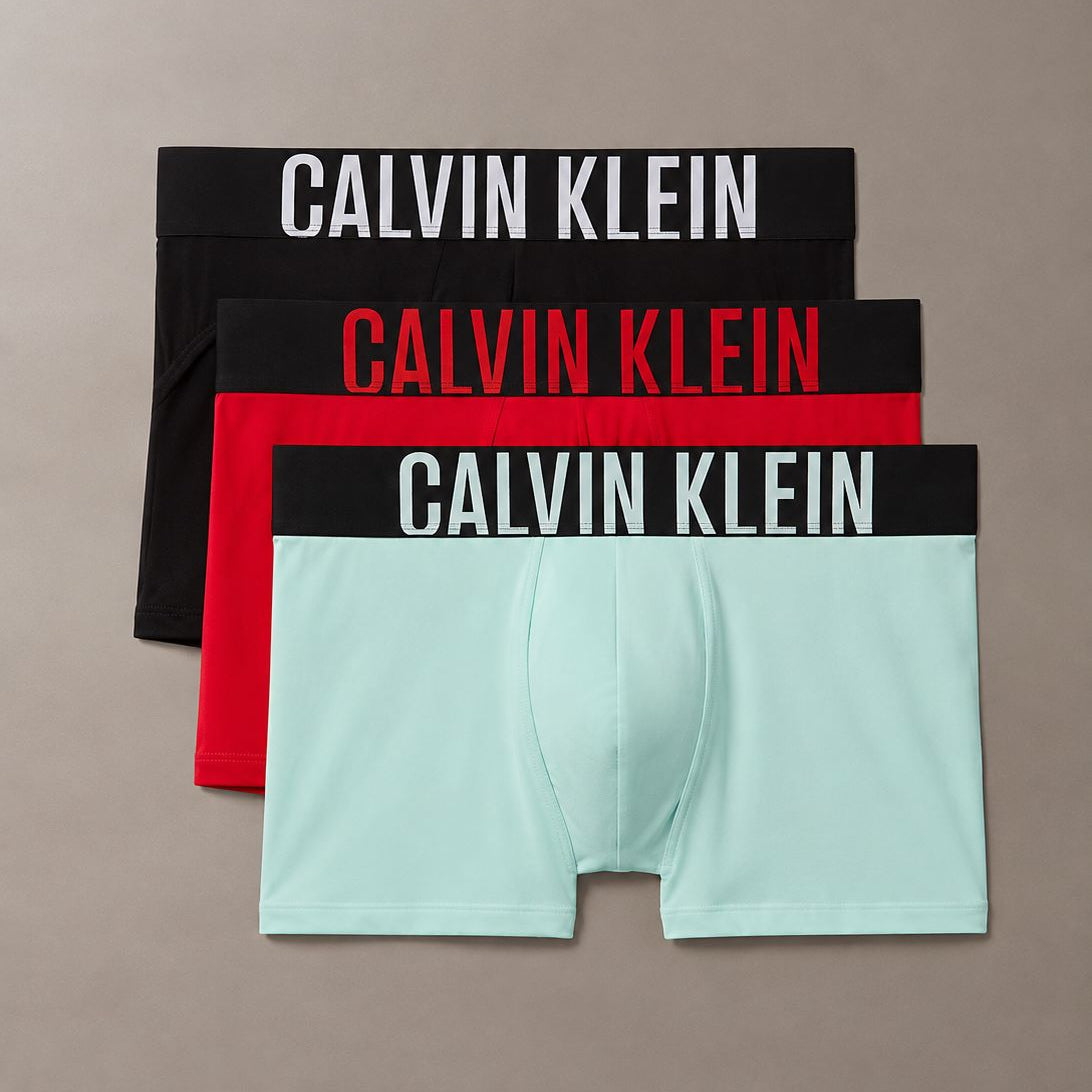 Three pairs of Calvin Klein boxer briefs stacked diagonally on a neutral taupe background. The top pair is black with white text, the middle is red with red text, and the bottom is mint green with a black waistband and white text.