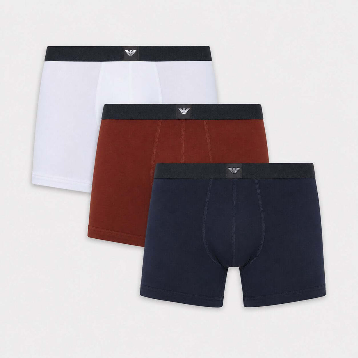 Three pairs of men's boxer briefs arranged in a fan layout on a light gray background: white, rust red, and navy blue. Each pair has a dark waistband with a small silver embroidered eagle logo at the center.