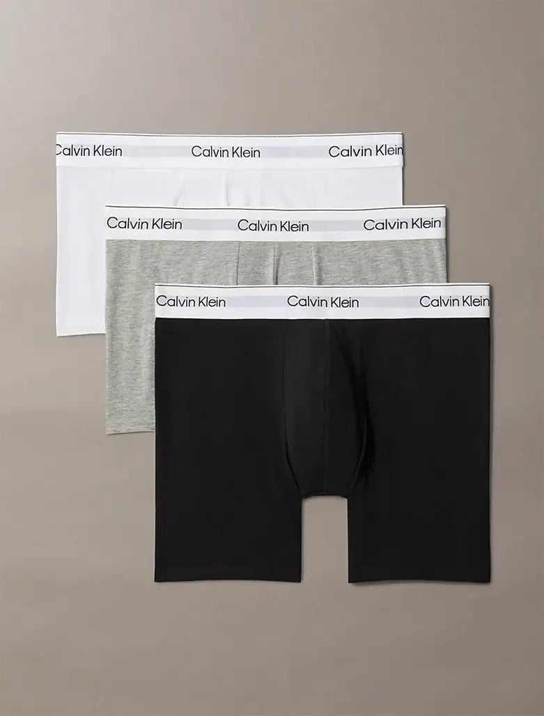 Three pairs of Calvin Klein boxer briefs in black, gray, and white on a beige background.
