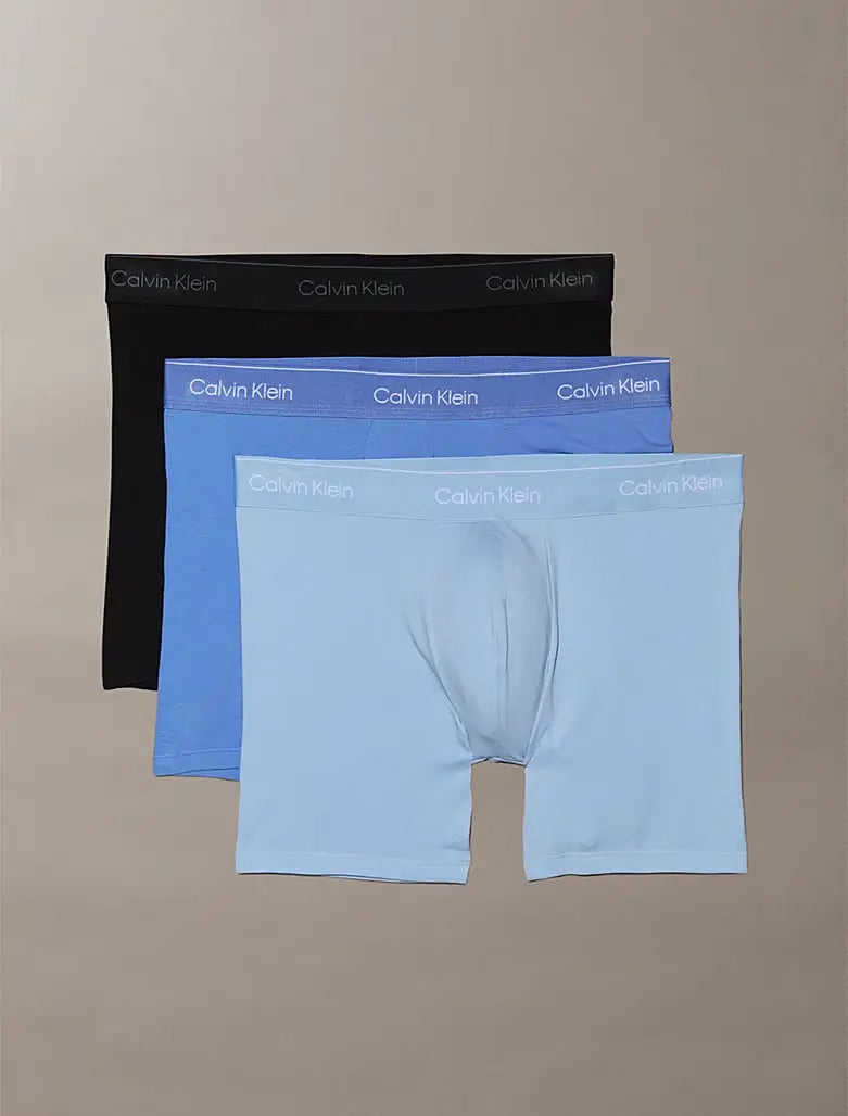 Three pairs of Calvin Klein boxer briefs in black, blue, and light blue on a beige background.