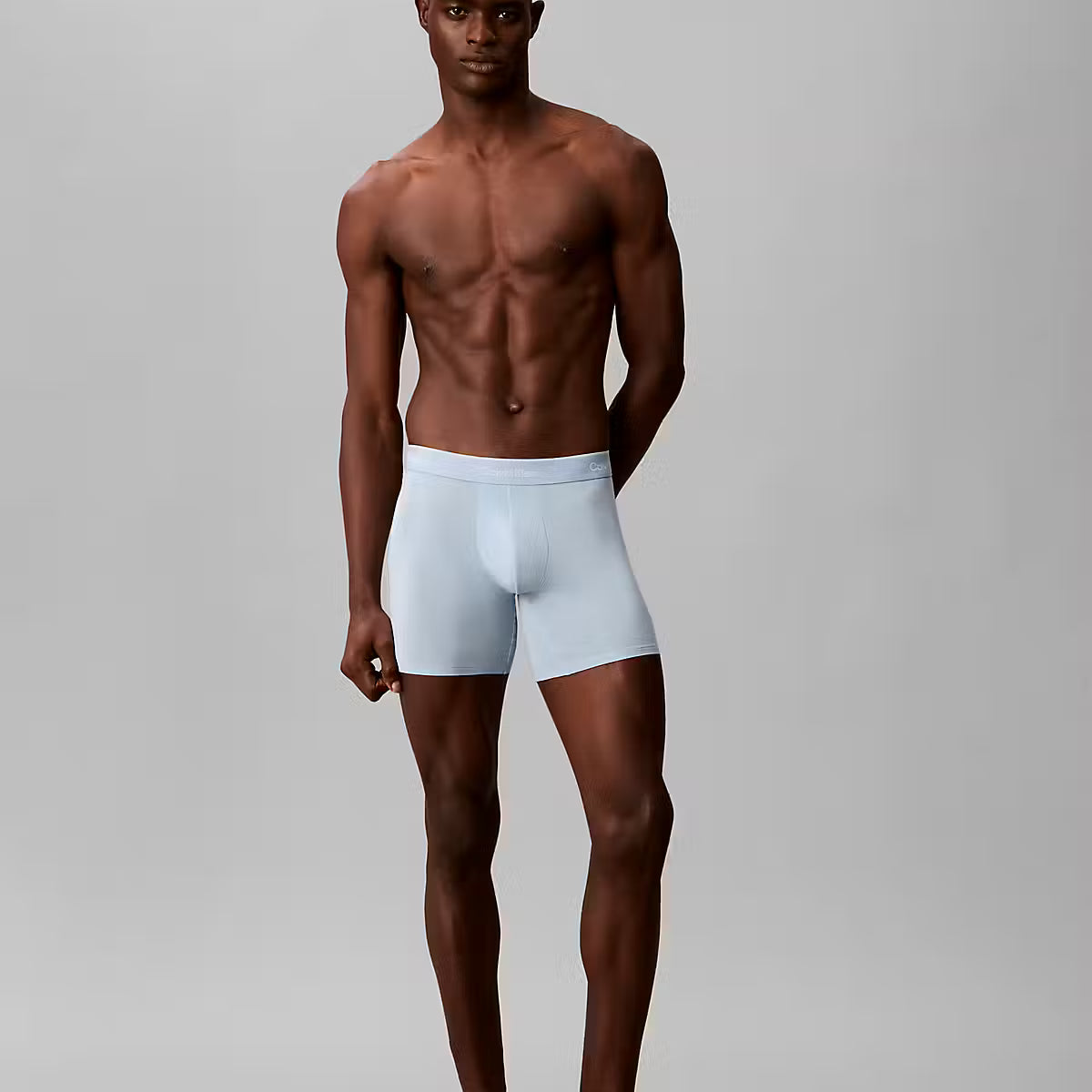 A shirtless male model stands against a light gray studio background wearing light blue boxer briefs.