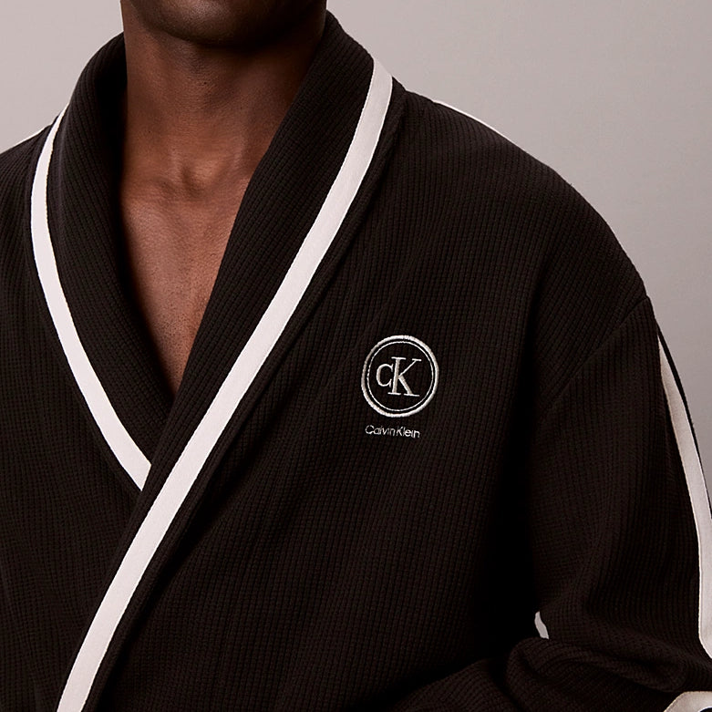A close-up of a man wearing a dark brown waffle-knit robe with white contrast trim and a white embroidered Calvin Klein logo on the chest.