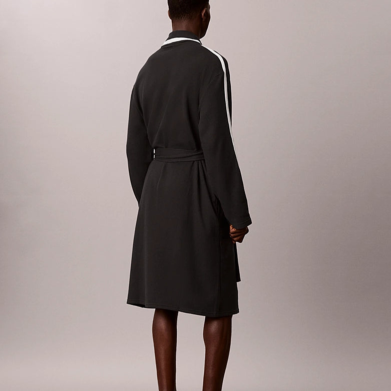 A man stands barefoot from the back wearing a dark charcoal gray robe with white stripes down the sleeves and a white collar, set against a plain light gray studio background.
