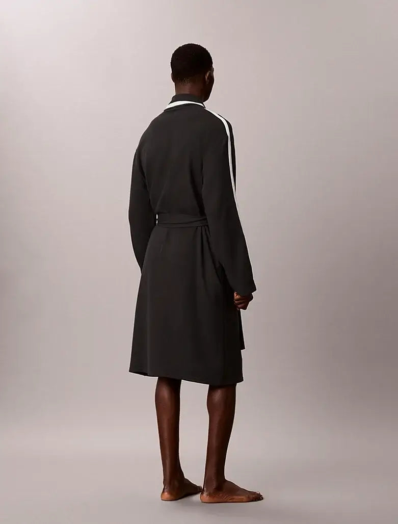 A man stands barefoot from the back wearing a dark charcoal gray robe with white stripes down the sleeves and a white collar, set against a plain light gray studio background.