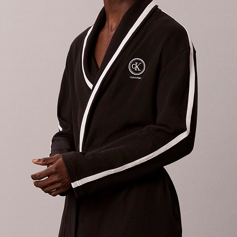 A man wearing a black ribbed cotton bathrobe with white contrast piping and a white embroidered Calvin Klein logo on the chest, set against a plain light gray studio background.