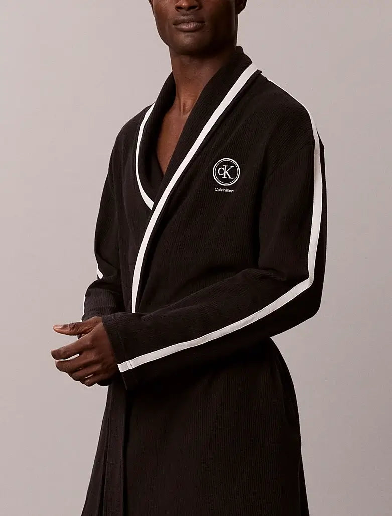 A man wearing a black ribbed cotton bathrobe with white contrast piping and a white embroidered Calvin Klein logo on the chest, set against a plain light gray studio background.