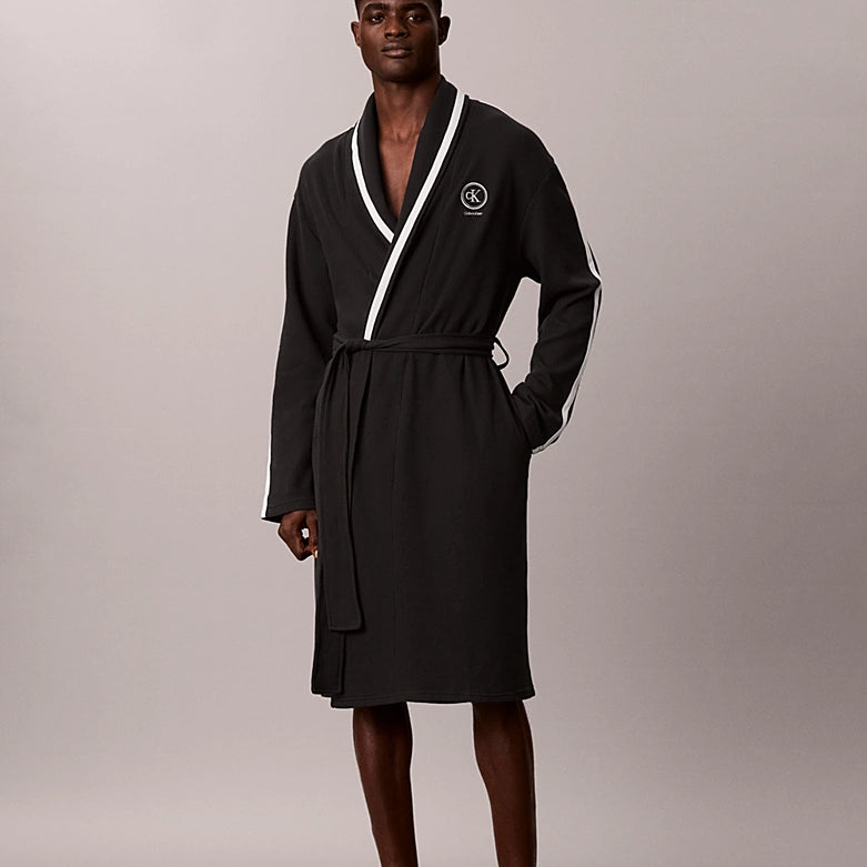 A man modeling a black bathrobe with white contrast piping and a white circular logo on the chest, standing against a neutral light gray studio background.