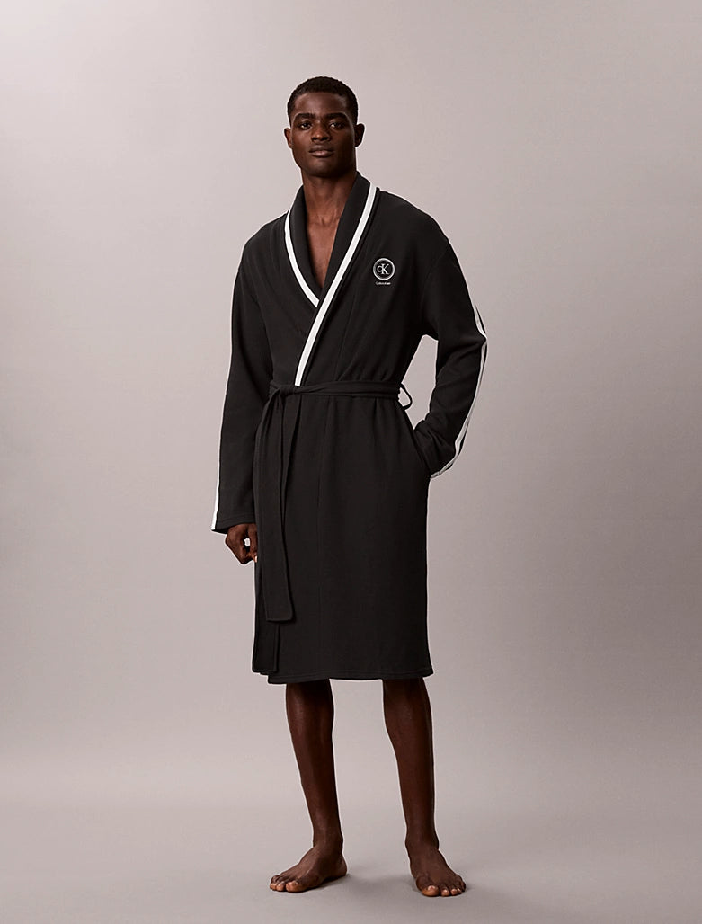 A man modeling a black bathrobe with white contrast piping and a white circular logo on the chest, standing against a neutral light gray studio background.