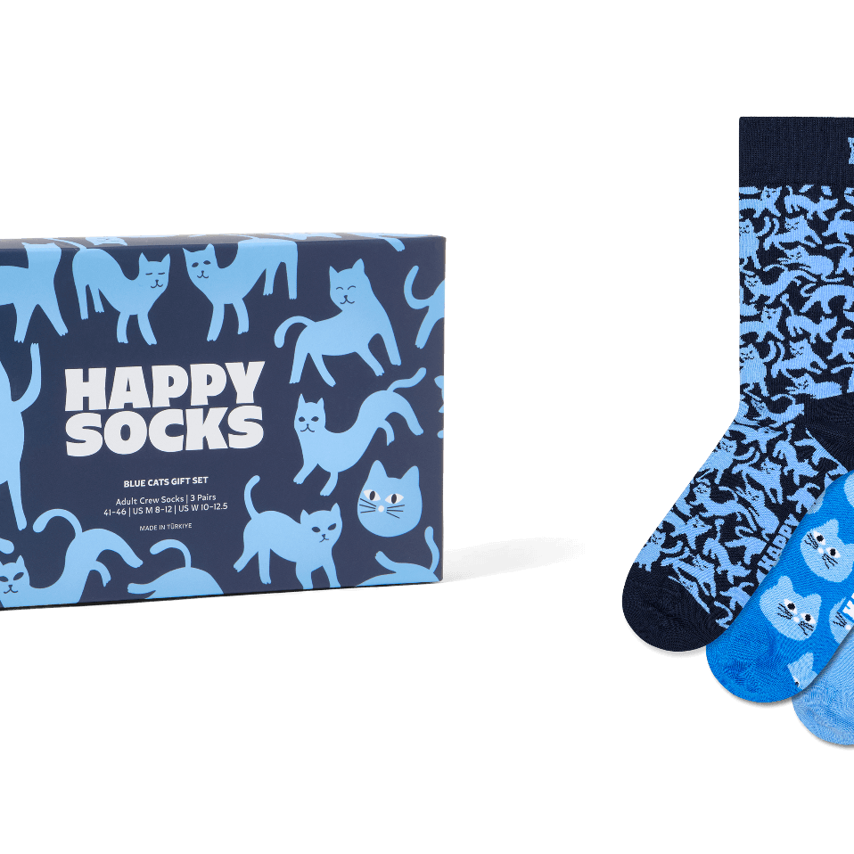 A boxed set of three patterned socks displayed beside the retail box on a light gray background. The packaging and sock designs feature blue cat illustrations and the text 'HAPPY SOCKS'.