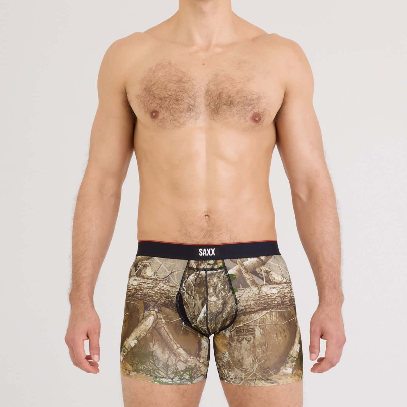 A man wearing camouflage boxer briefs with a black waistband featuring the SAXX logo, photographed against a light gray studio background.
