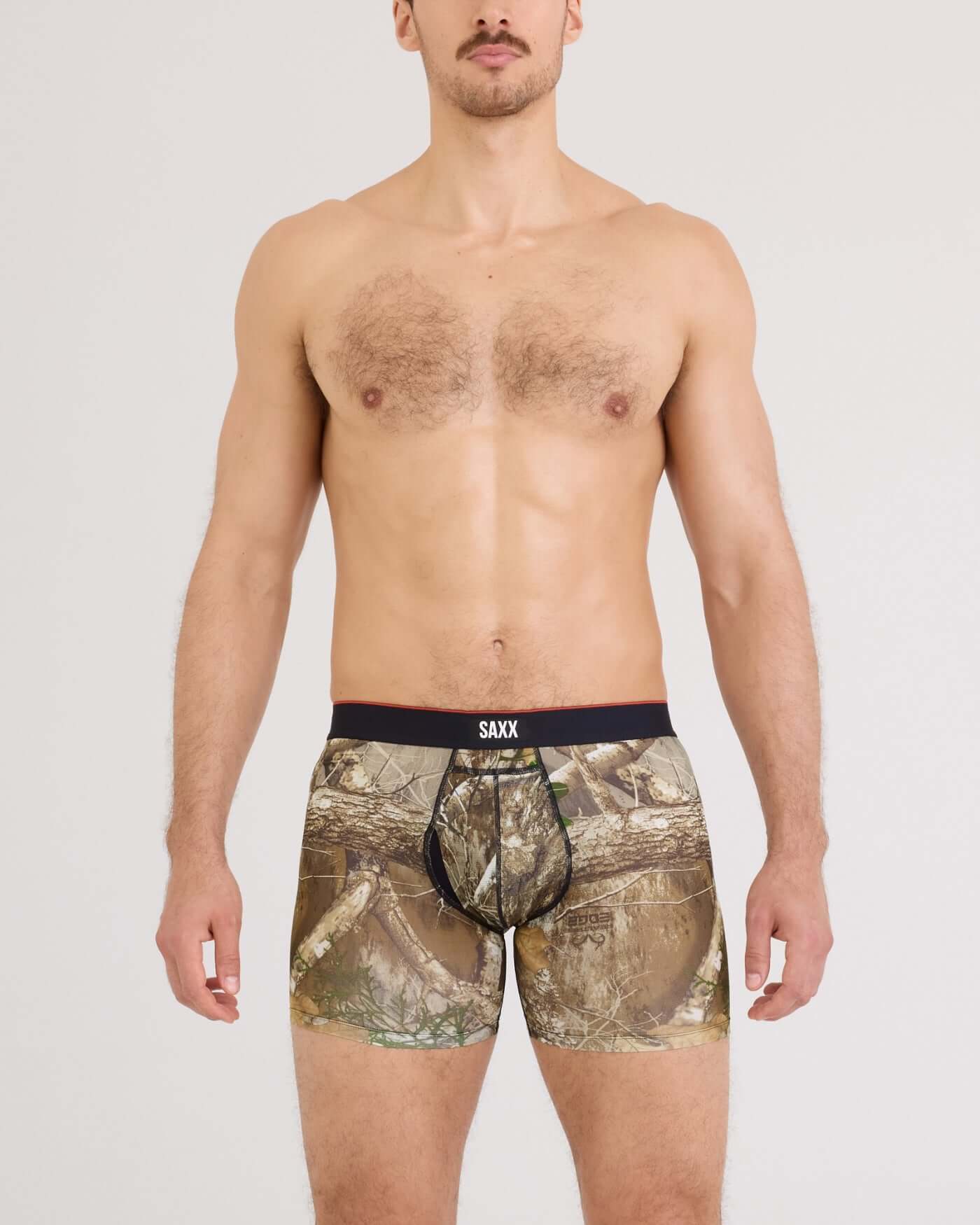 A man wearing camouflage boxer briefs with a black waistband featuring the SAXX logo, photographed against a light gray studio background.