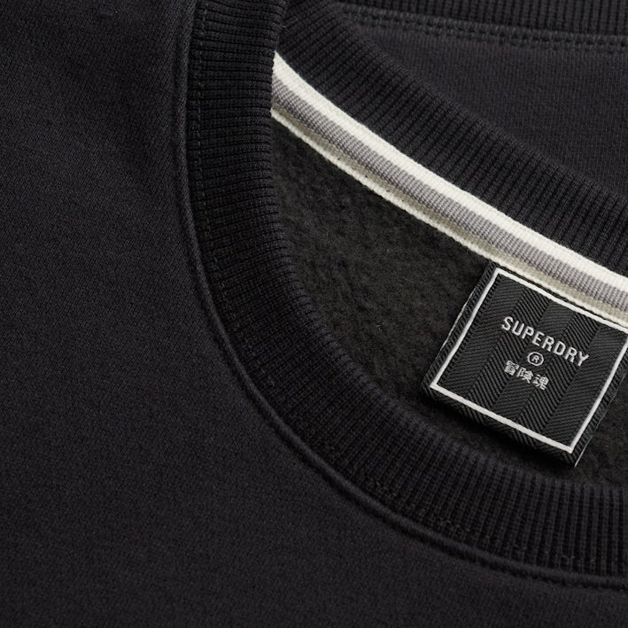 Close-up of a black crewneck sweatshirt showing the ribbed collar, a white stripe detail inside the neckline, and a black Superdry brand label.