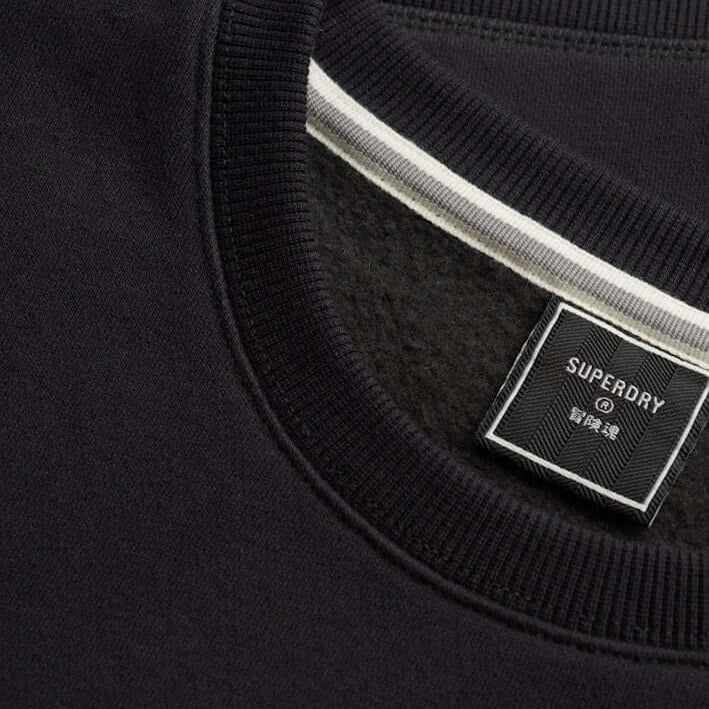 Close-up of a black crewneck sweatshirt showing the ribbed collar, a white stripe detail inside the neckline, and a black Superdry brand label.