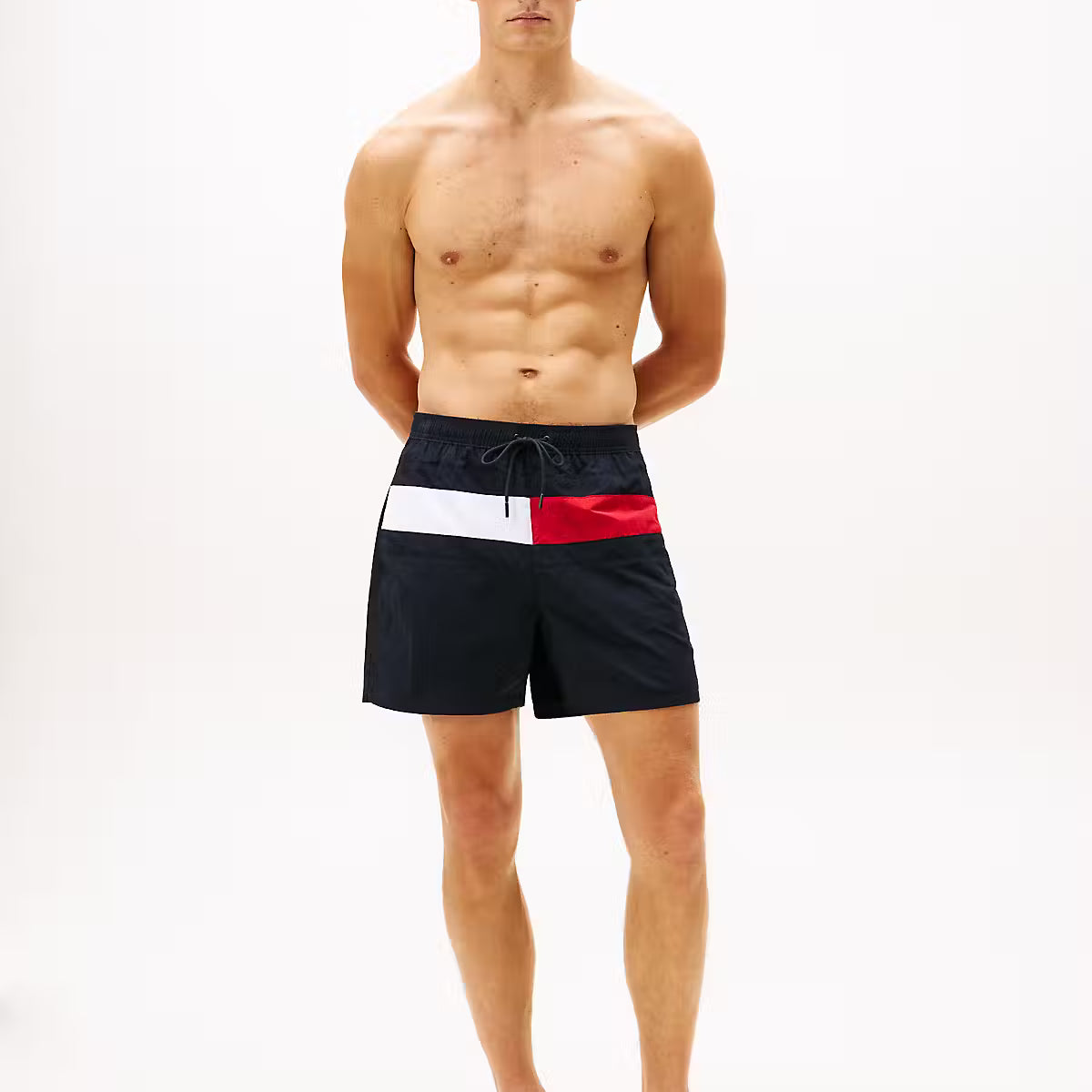 A shirtless male model stands against a plain white studio background wearing navy swim shorts with a white and red horizontal stripe detail at the waist.