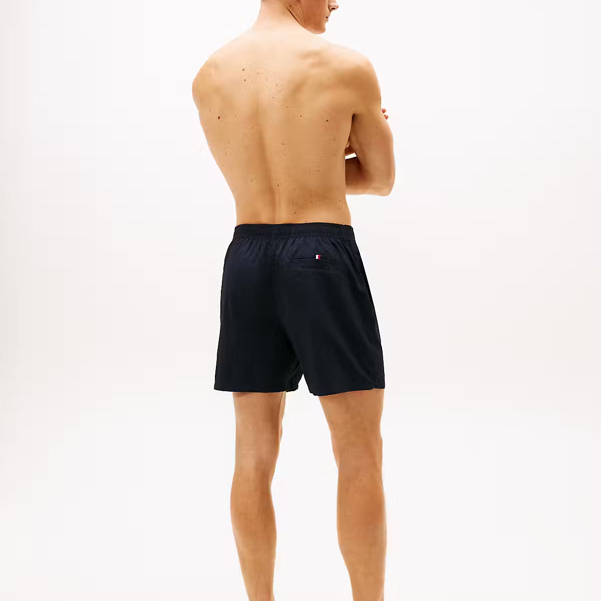 A man shown from the back wearing dark navy blue swim shorts, standing against a plain white studio background.