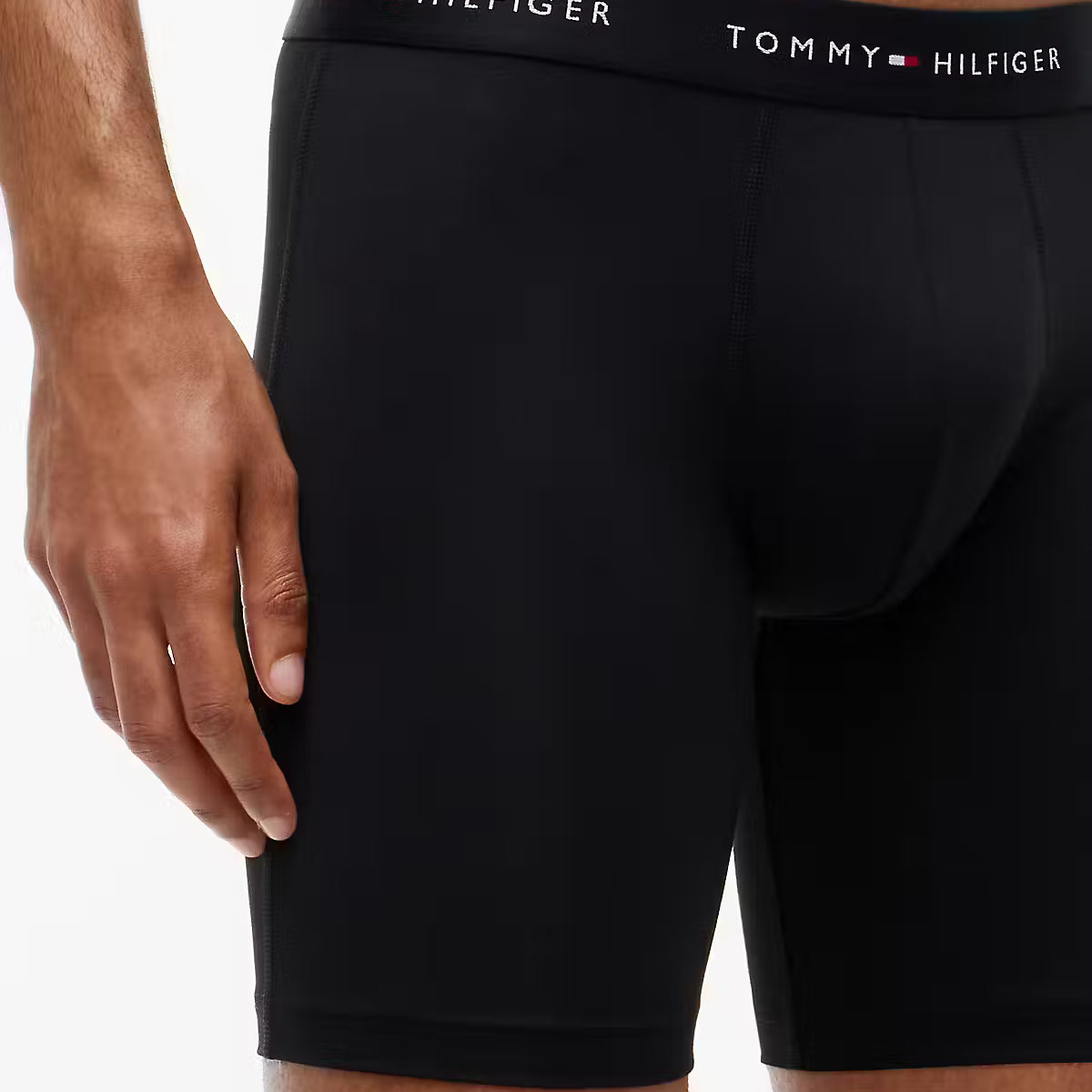 A close-up of a model wearing black Tommy Hilfiger boxer briefs, showing the branded elastic waistband and the fit of the underwear.