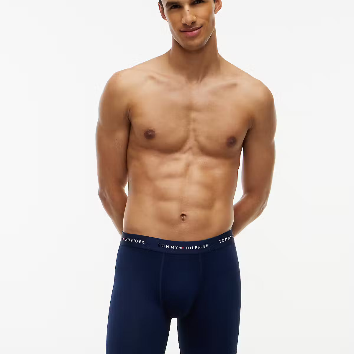 A shirtless male model wearing Tommy Hilfiger navy blue boxer briefs, standing against a plain white studio background.