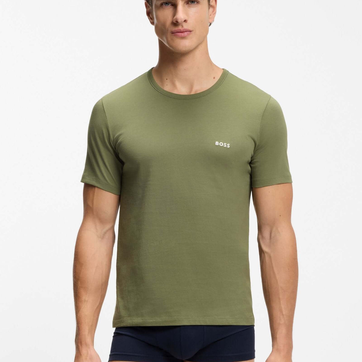 A male model wearing an olive green short-sleeve crewneck t-shirt with a small white BOSS logo on the left chest, paired with navy blue boxer briefs, against a plain light gray studio background.