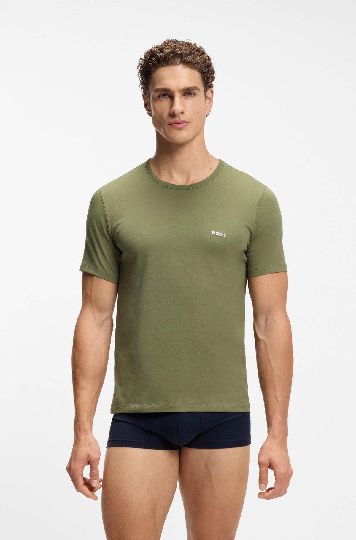 A male model wearing an olive green short-sleeve crewneck t-shirt with a small white BOSS logo on the left chest, paired with navy blue boxer briefs, against a plain light gray studio background.