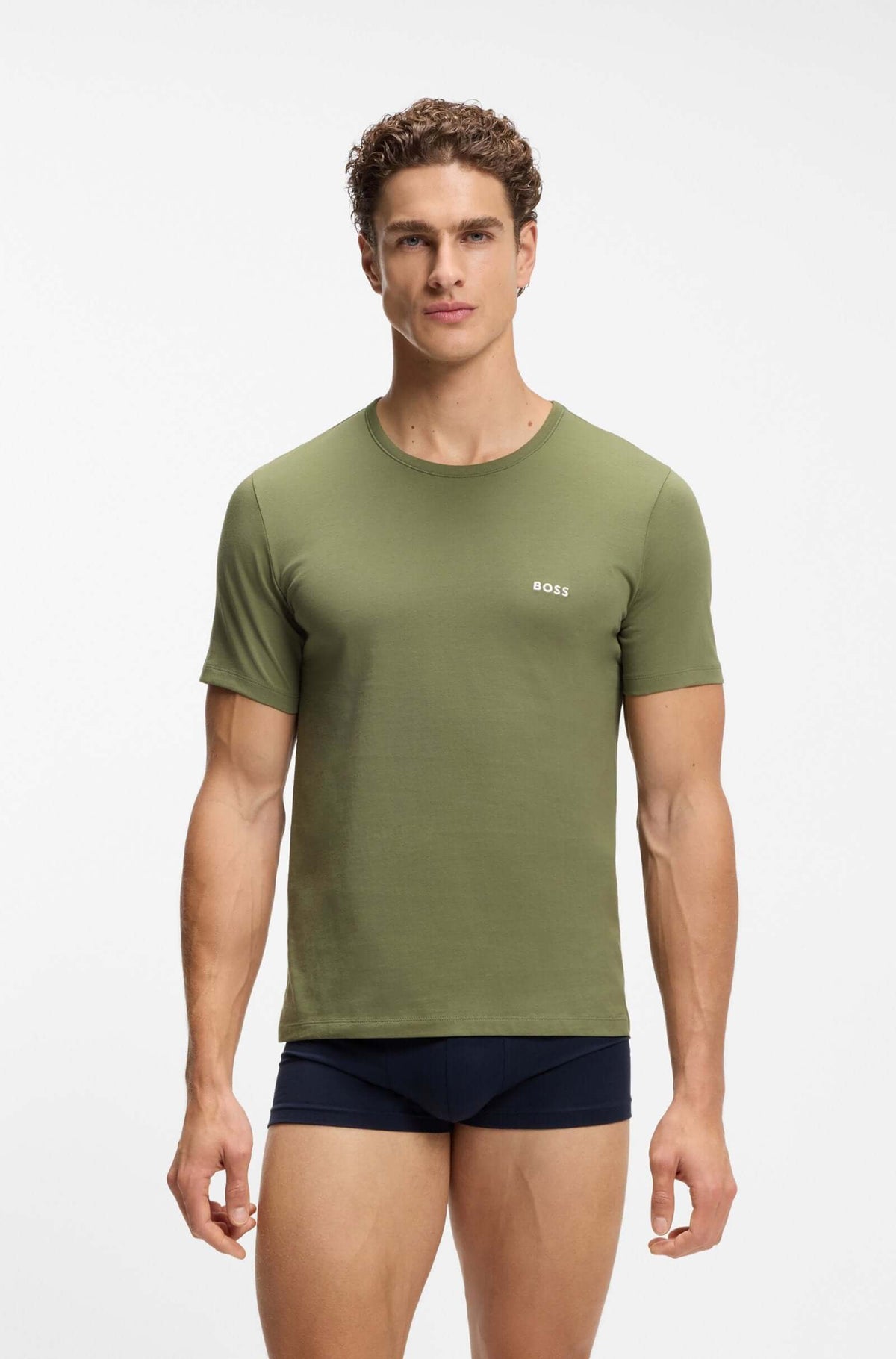 A male model wearing an olive green short-sleeve crewneck t-shirt with a small white BOSS logo on the left chest, paired with navy blue boxer briefs, against a plain light gray studio background.
