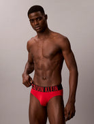 A shirtless male model wearing red Calvin Klein briefs with a black waistband and red logo text, photographed against a plain light gray studio background.