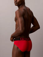 A man shown from the back wearing bright red Calvin Klein briefs with a black waistband and red logo text, against a plain light gray studio background.