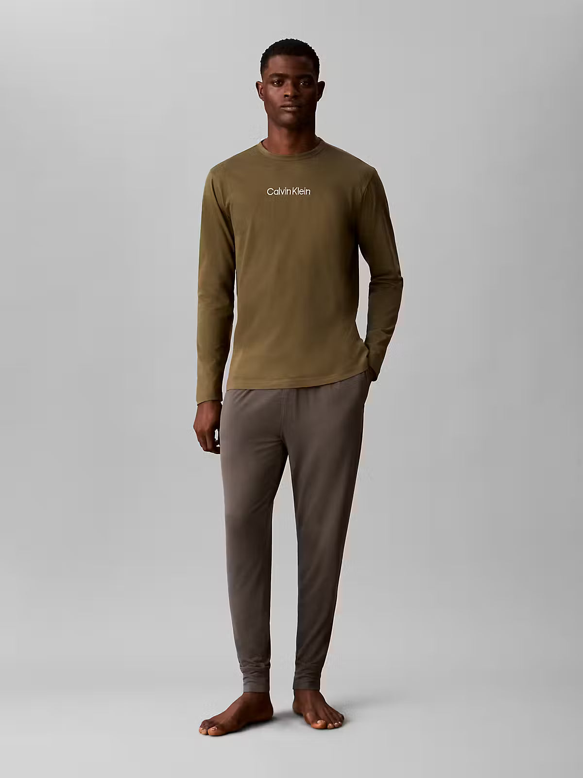 A man modeling a two-piece Calvin Klein loungewear set consisting of an olive green long-sleeve shirt and dark brown pants, standing against a neutral gray studio background.