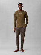 A man modeling a two-piece Calvin Klein loungewear set consisting of an olive green long-sleeve shirt and dark brown pants, standing against a neutral gray studio background.
