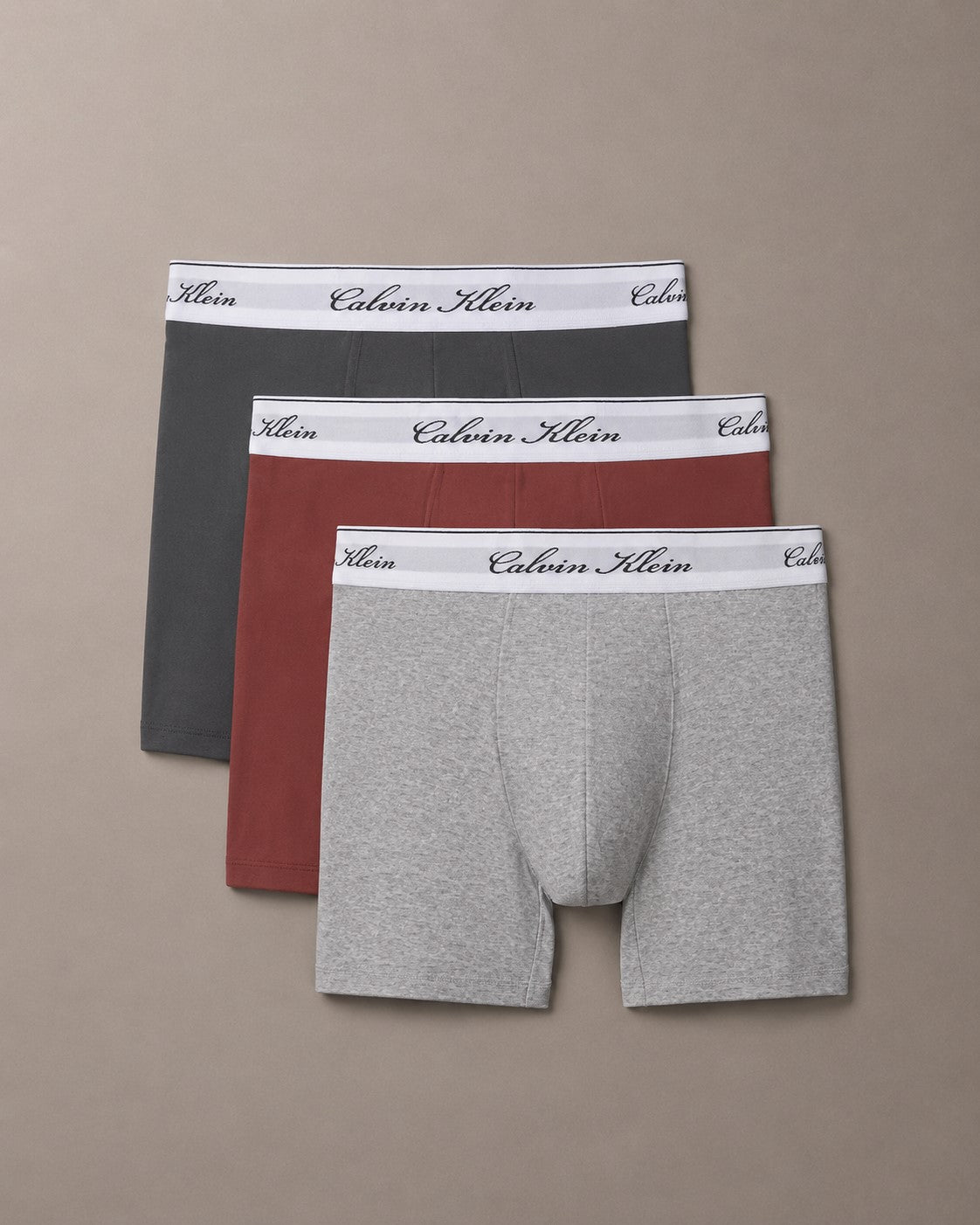 Three pairs of Calvin Klein men's boxer briefs in dark gray, rust red, and light heather gray, arranged in a fan shape on a solid taupe background.