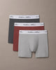 Three pairs of Calvin Klein men's boxer briefs in dark gray, rust red, and light heather gray, arranged in a fan shape on a solid taupe background.