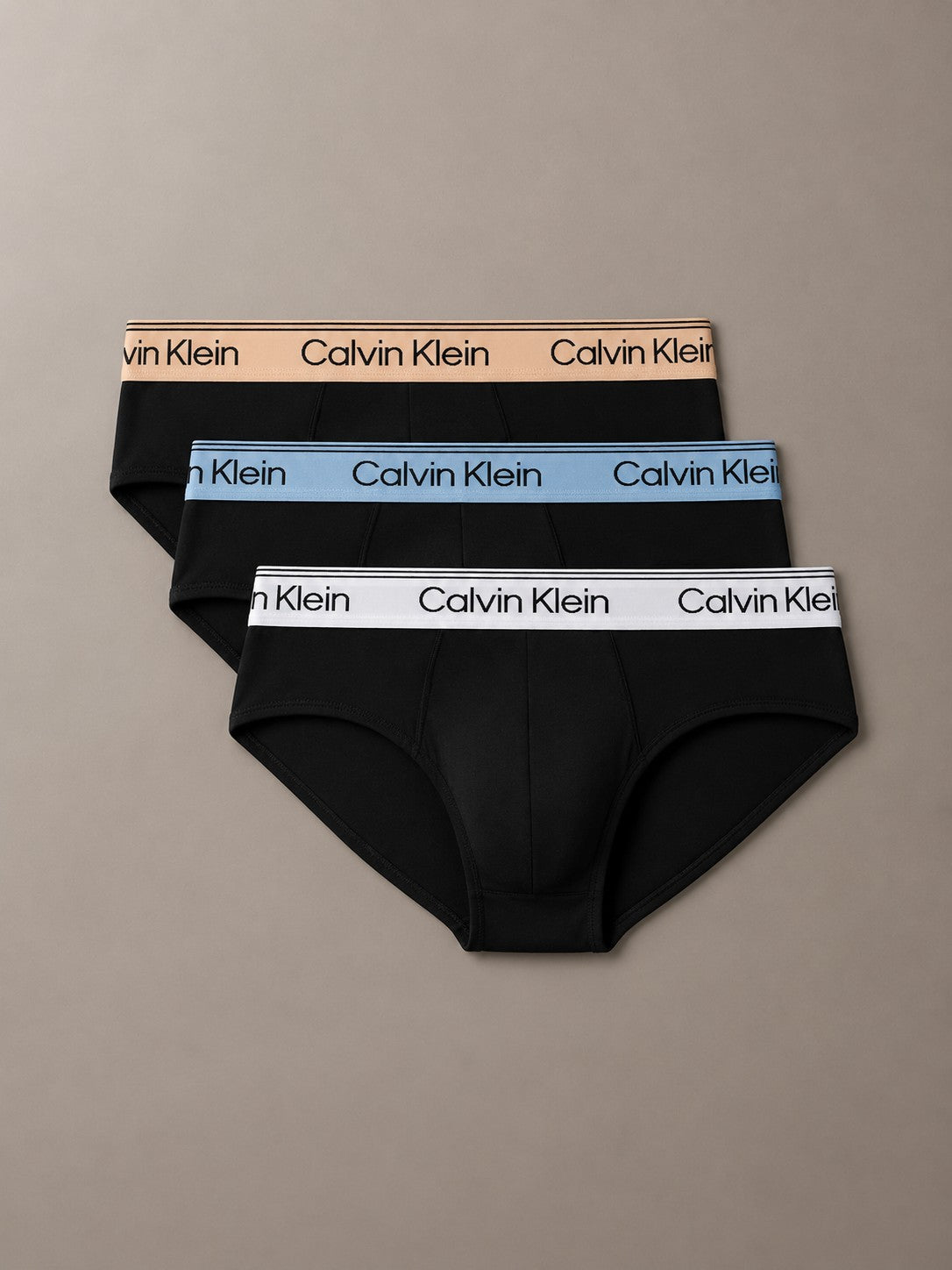 Three pairs of Calvin Klein men's briefs stacked in a fan arrangement on a neutral taupe background. The briefs are black with contrasting elastic waistbands in beige, light blue, and white, each featuring the repeating Calvin Klein logo.