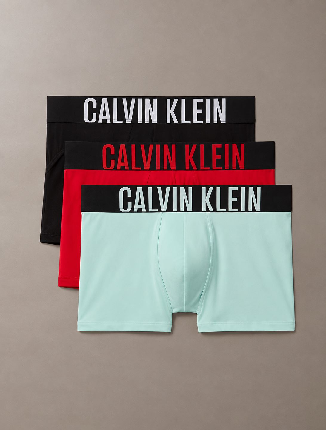 Three pairs of Calvin Klein boxer briefs stacked diagonally on a neutral taupe background. The top pair is black with white text, the middle is red with red text, and the bottom is mint green with a black waistband and white text.
