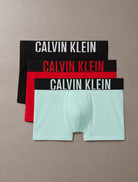 Three pairs of Calvin Klein boxer briefs stacked diagonally on a neutral taupe background. The top pair is black with white text, the middle is red with red text, and the bottom is mint green with a black waistband and white text.