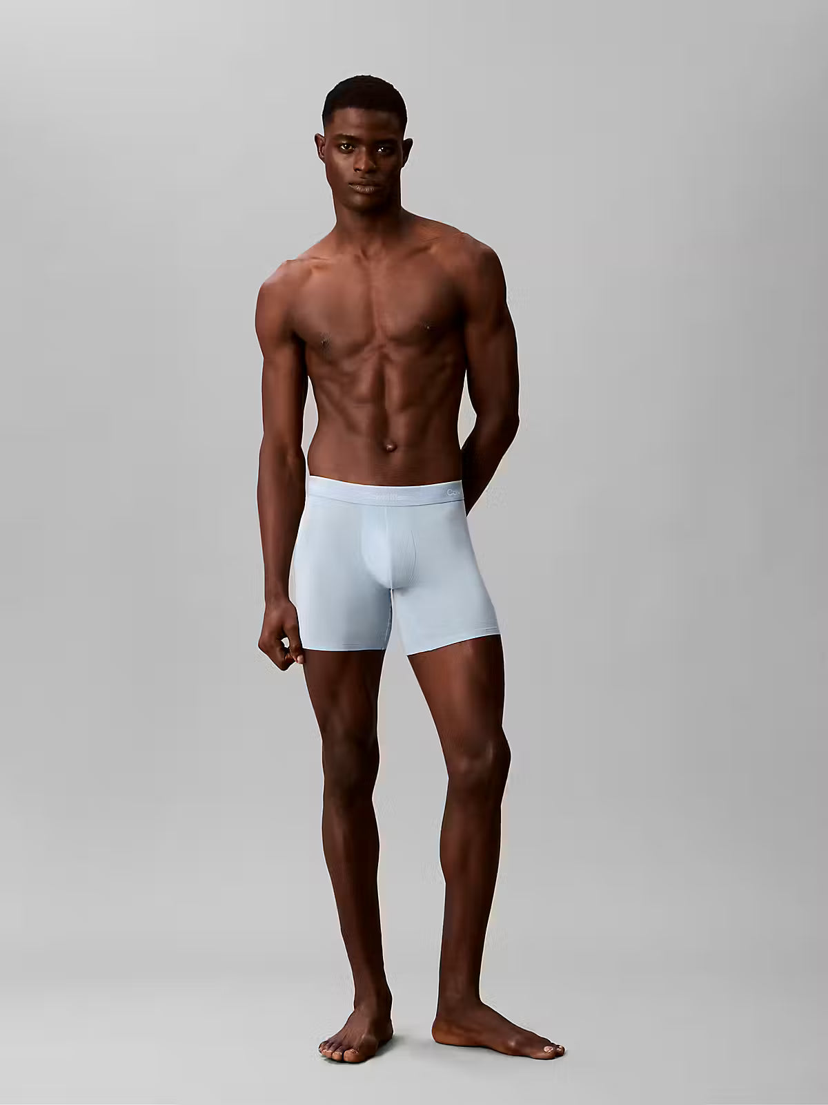 A shirtless male model stands against a light gray studio background wearing light blue boxer briefs.