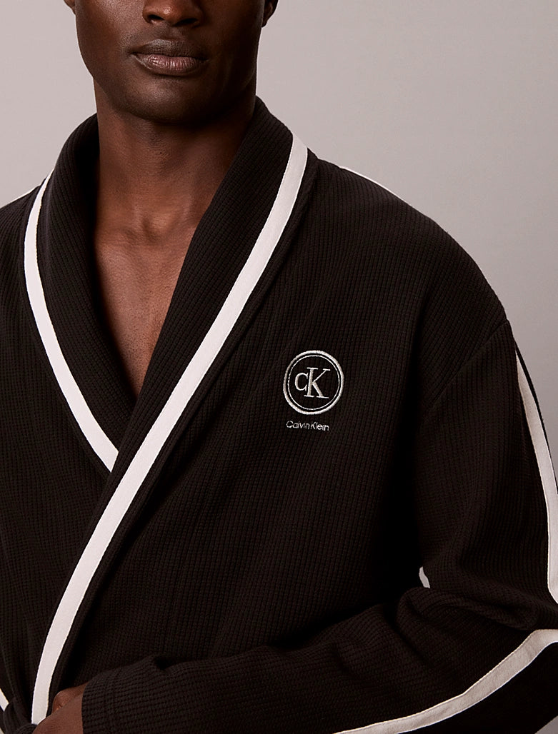 A close-up of a man wearing a dark brown waffle-knit robe with white contrast trim and a white embroidered Calvin Klein logo on the chest.