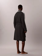 A man stands barefoot from the back wearing a dark charcoal gray robe with white stripes down the sleeves and a white collar, set against a plain light gray studio background.