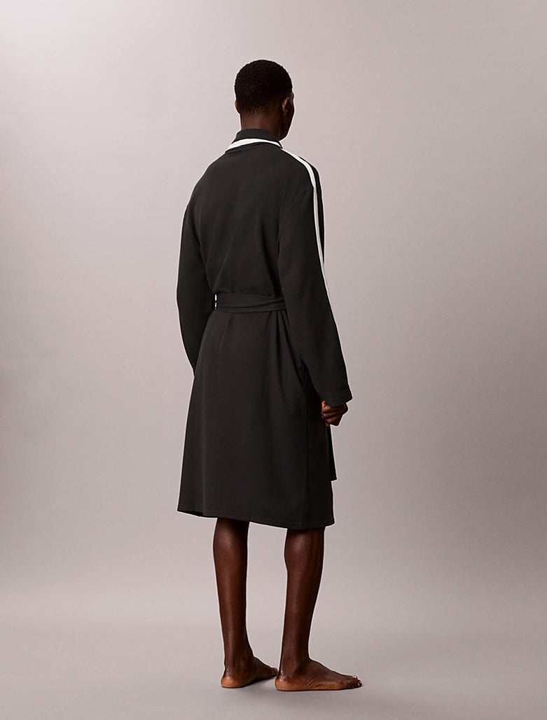 A man stands barefoot from the back wearing a dark charcoal gray robe with white stripes down the sleeves and a white collar, set against a plain light gray studio background.