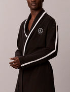 A man wearing a black ribbed cotton bathrobe with white contrast piping and a white embroidered Calvin Klein logo on the chest, set against a plain light gray studio background.