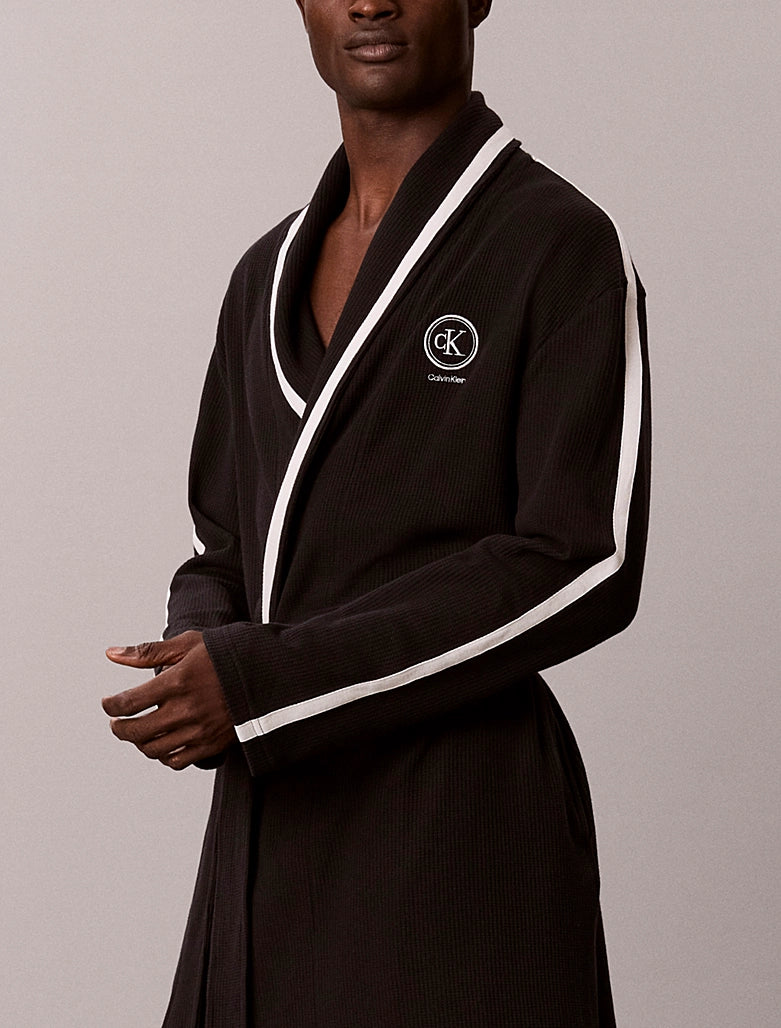 A man wearing a black ribbed cotton bathrobe with white contrast piping and a white embroidered Calvin Klein logo on the chest, set against a plain light gray studio background.
