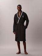 A man modeling a black bathrobe with white contrast piping and a white circular logo on the chest, standing against a neutral light gray studio background.