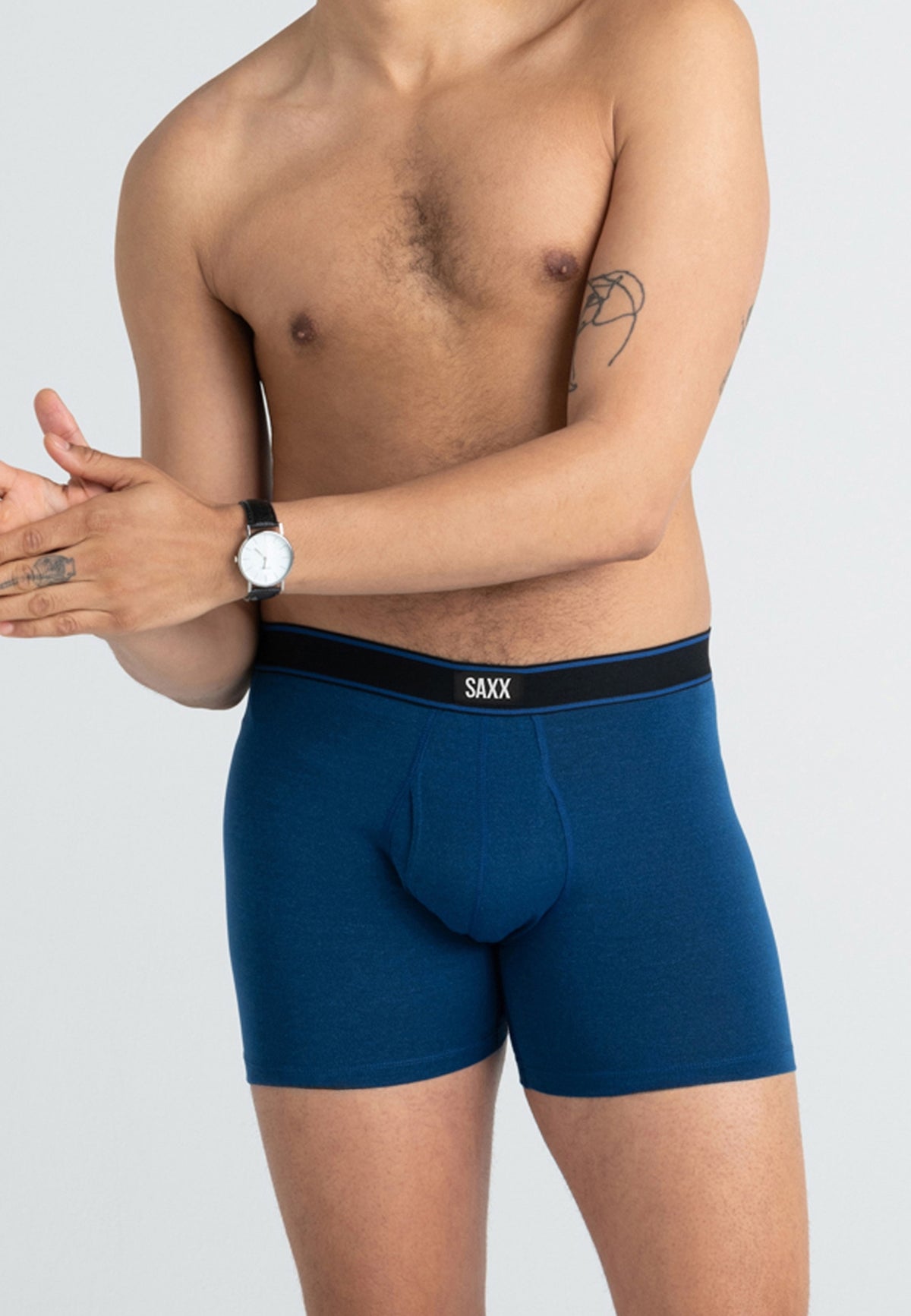 A man wearing blue boxer briefs with a black waistband featuring the brand name 'SAXX' in white, shown from the chest down against a light gray background.