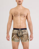 A man wearing camouflage boxer briefs with a black waistband featuring the SAXX logo, photographed against a light gray studio background.
